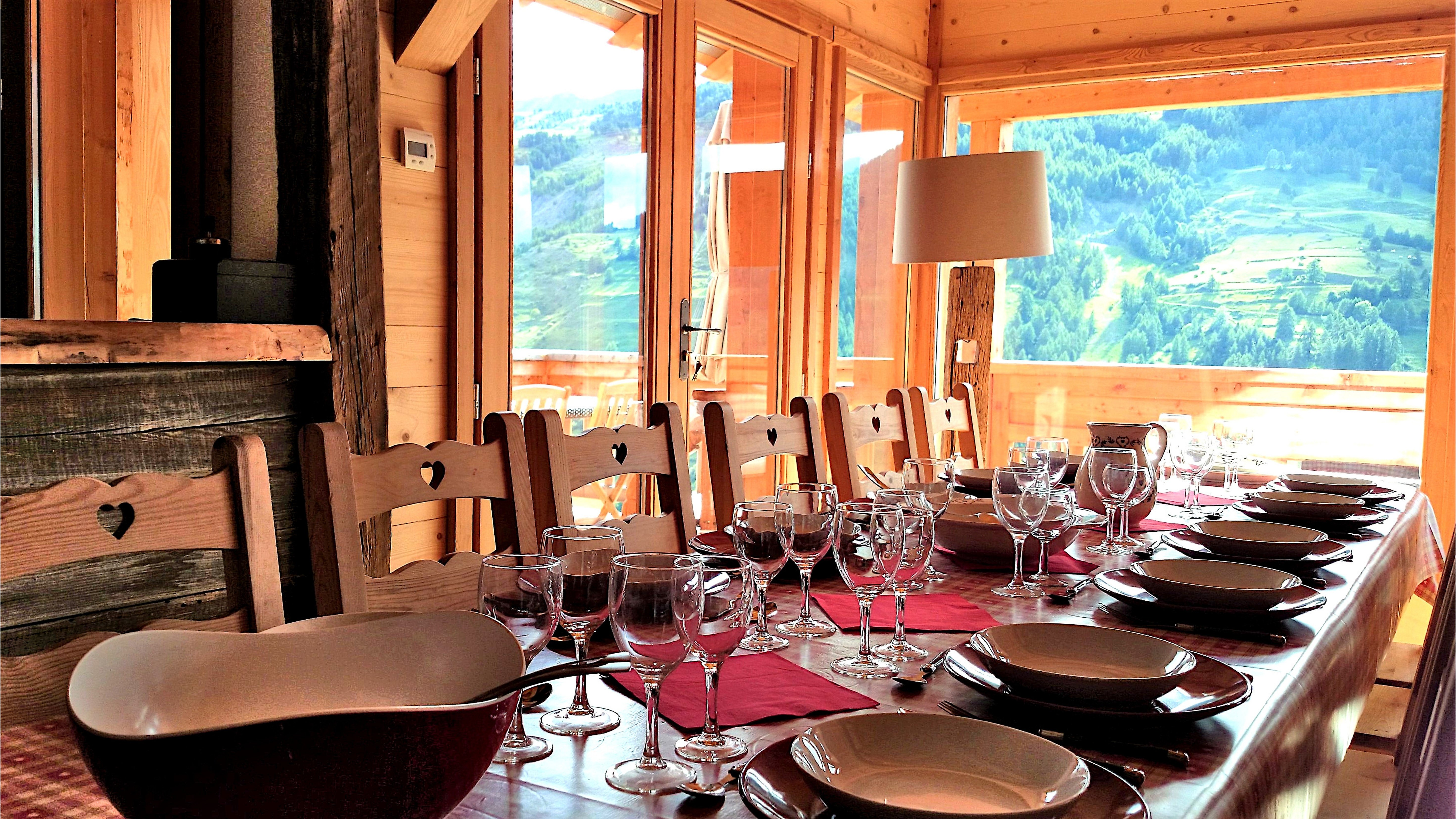 Grande table intérieure dressée pour un repas de 12 personnes au Chalet B à Vars, installée devant une grande fenêtre ouverte sur la montagne.